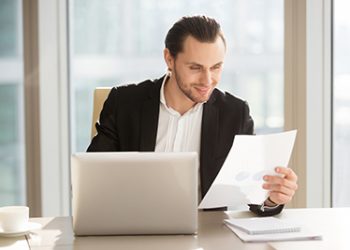Young guy satisfied with company financial results, businessman enjoys good rate of growth. Entrepreneur looking on document with graphs and diagrams at desk in front of laptop. Manager reading notice